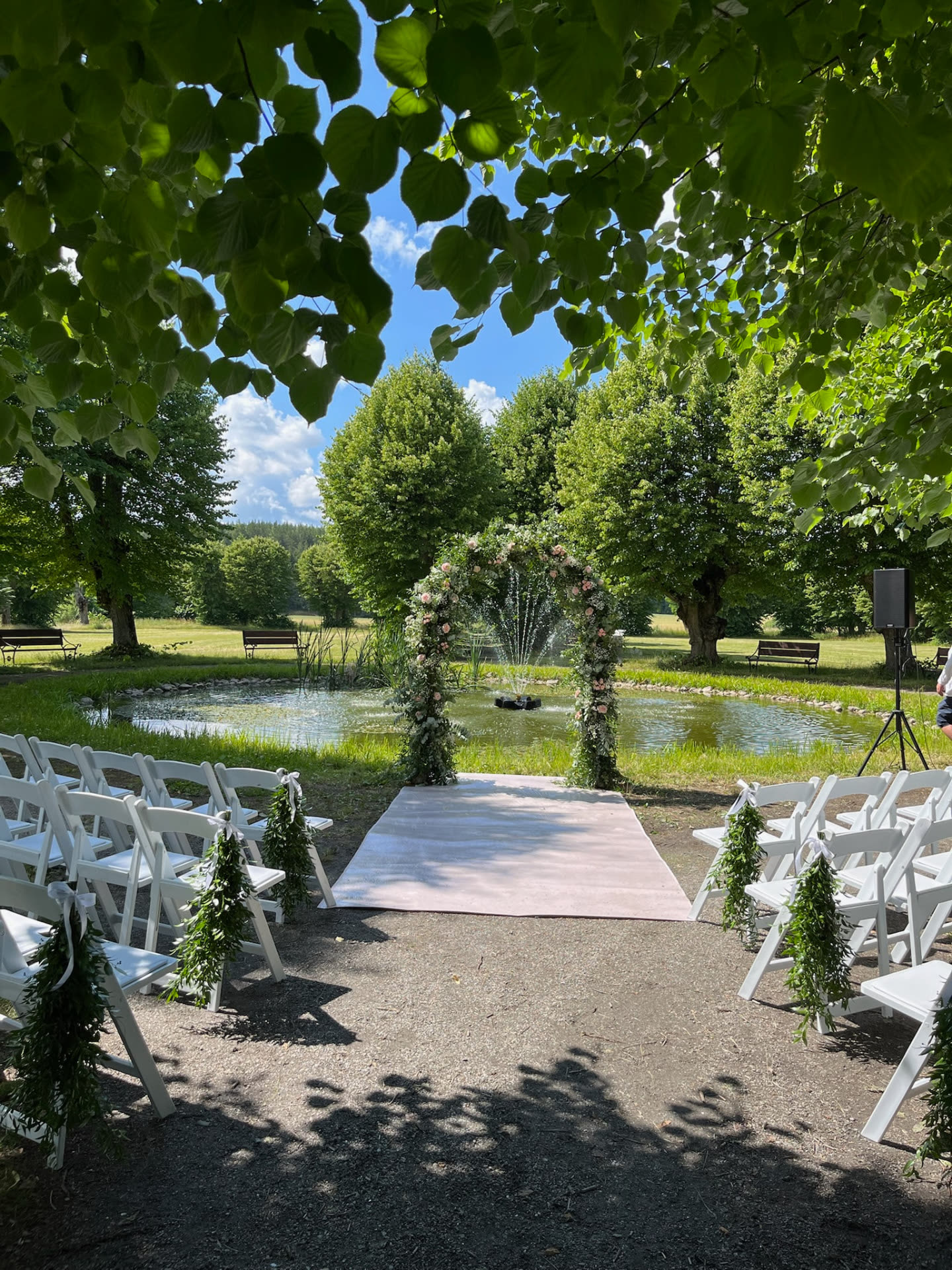 Vigsel vid dammen med blomsterbåge, matta och vita stolar